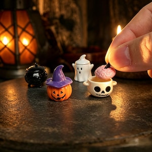 May include: A set of miniature ceramic Halloween figurines including a black cauldron, an orange pumpkin wearing a purple witch hat, a white ghost, and a white skull bowl with a pink brain-shaped lid being held by a hand.