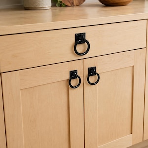 Könnte beinhalten: Heller Holzschrank mit drei schwarzen Metallringgriffen. Ein Ringgriff ist mittig an der oberen Schublade angebracht, während zwei passende Ringgriffe an den unteren Schranktüren befestigt sind. Die Beschläge haben eine quadratische Platte.