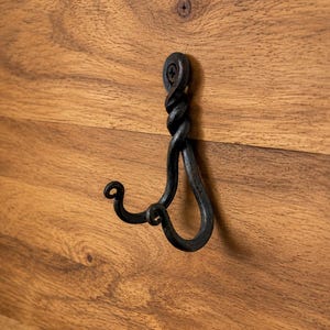 May include: A dark, hand-forged metal wall hook featuring a twisted vertical stem and two curved prongs. The hook is secured to a wooden surface with a single visible screw at the top, showcasing a textured, blackened iron finish throughout.