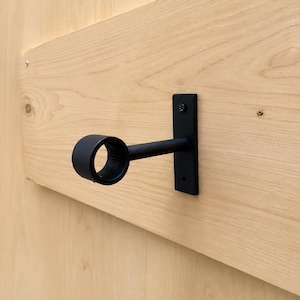 May include: A matte black metal curtain rod bracket featuring a rectangular wall-mounting plate with two screw holes and a cylindrical arm extending outwards to a circular ring designed to hold a curtain rod securely in place.
