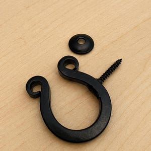 May include: A matte black metal hook with a curved, open-loop design and a pointed threaded screw end, accompanied by a separate small, circular black metal washer. The hook features a decorative curled tip at the end of the loop.