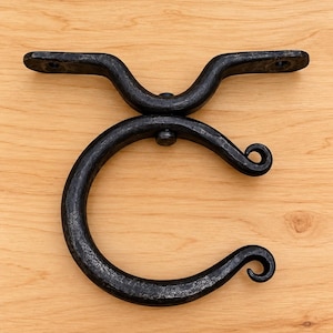 Op de afbeelding: Een zwarte, handgesmede metalen wandhaak met een cirkelvormig ringontwerp en een getextureerde, gehamerde afwerking. De haak bevat een horizontale montagebeugel met twee voorgeboorde schroefgaten aan de uiteinden.