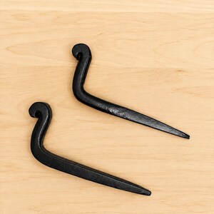Op de afbeelding: Twee zwarte metalen haken met een handgesmede uitstraling. Elke haak heeft een gebogen, gekruld uiteinde aan de bovenkant en een recht, taps toelopend lichaam dat smaller wordt naar de punt toe. Het metaaloppervlak heeft een matte, textuurafwerking.
