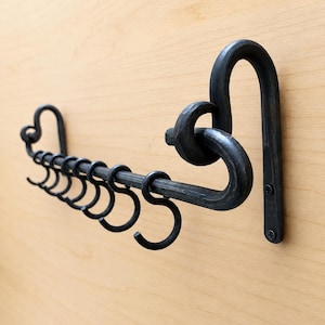 May include: A black forged metal wall-mounted rack featuring a horizontal bar with multiple sliding S-shaped hooks. The rack is secured to the wall with two visible screws at the end, showcasing a dark, textured finish and a sturdy, industrial design.