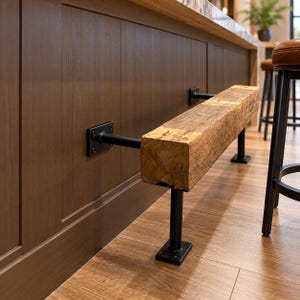 Puede incluir: Un reposapiés que consiste en una pieza gruesa y rectangular de madera recuperada montada en una isla de cocina con tuberías de metal negro y soportes de pared cuadrados. La viga de madera presenta una superficie rugosa con vetas visibles.