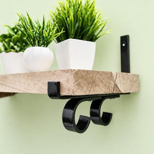 May include: A wall-mounted shelf featuring a thick, light-toned natural wood plank supported by two black metal brackets with a curved, scroll-like design. Three small white pots containing artificial green grass sit on the shelf surface.
