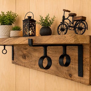 Puede incluir: Un estante de pared de madera con acabado de veta natural y soportes de metal negro con ganchos circulares integrados. El estante sostiene dos plantas verdes en macetas, una linterna de metal negro con luz interior y un modelo de bicicleta.
