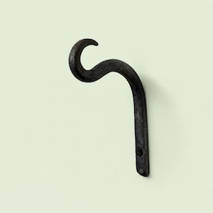 Op de afbeelding: Een donkergrijze, gebogen metalen wandhaak met een getextureerde, matte afwerking. De haak heeft een opvallend S-vormig silhouet dat taps toeloopt naar het bovenste uiteinde. Een zwarte schroef is zichtbaar bij het onderste bevestigingspunt.