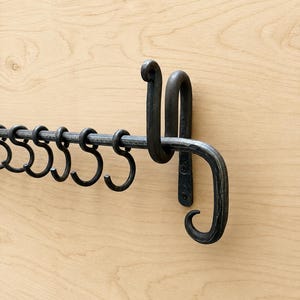 Op de afbeelding: Een aan de muur gemonteerd rek van donkergrijs metaal met een horizontale stang die wordt ondersteund door twee gebogen beugels. Een reeks bijpassende S-vormige haken hangt aan de stang, elk ontworpen om langs de stang te schuiven voor ophangen.