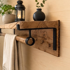 Puede incluir: Un estante de madera montado en la pared con un acabado de grano natural y un sistema de soportes de metal negro. La unidad incluye una barra horizontal de madera sostenida por anillos metálicos circulares, con una toalla de tela beige colgada.