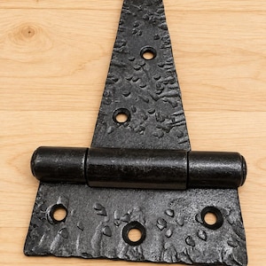 Puede incluir: Una bisagra en T negra con una superficie de metal texturizada y martillada. La bisagra incluye una placa de montaje triangular con tres orificios circulares para tornillos y una placa base rectangular con tres orificios adicionales para su instalación.
