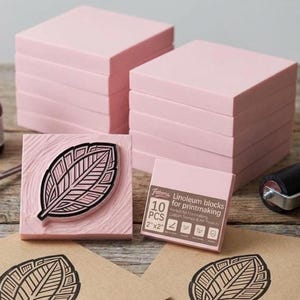 May include: A set of square pink linoleum blocks for printmaking, including a block carved with a leaf design. The packaging text reads Fittoria Linoleum blocks for printmaking, 10 PCS, 2"x2". The blocks are stacked in two piles on a wooden surface.