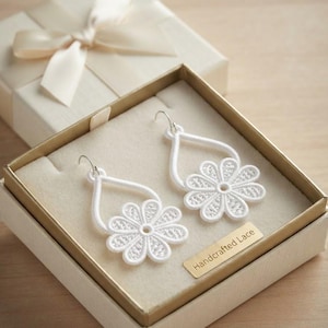 May include: A pair of white lace earrings featuring a teardrop shape with a flower motif, presented in a square gift box with a cream ribbon and a gold-toned plaque that reads Handcrafted Lace. The earrings have silver-toned metal hooks.