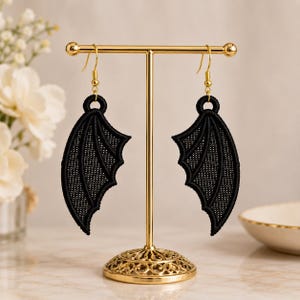May include: A pair of black embroidered bat wing earrings hanging from a gold-toned metal T-bar display stand. The earrings feature a detailed stitched texture and are suspended by gold-colored fishhook wires, showcasing a symmetrical design.