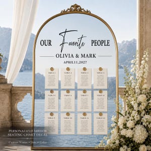 May include: An arched mirror with a gold frame featuring black text that reads OUR FAVORITE PEOPLE, OLIVIA & MARK, and APRIL 11, 2027. Twelve white rectangular cards are attached to the mirror, each labeled TABLE 1 through 12 with gold wax seals.
