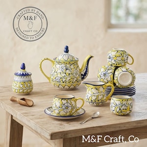 Pode incluir: Um conjunto de chá em cerâmica com padrão floral amarelo e branco com detalhes em azul. O conjunto inclui um bule com tampa, um açucareiro com tampa, uma leiteira, quatro chávenas e pires. Uma pequena colher de madeira e uma de metal estão presentes.