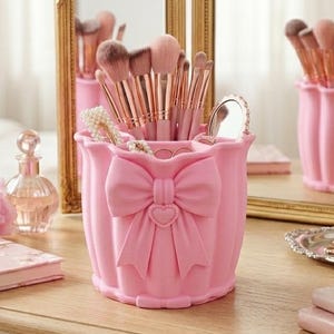Op de afbeelding: Een lichtroze make-up kwastenhouder met een opvallende strik en hartdetail aan de voorzijde. De cilindervormige houder heeft een gegolfde rand en is gevuld met diverse make-up kwasten met roségouden handvatten en zachte haren.