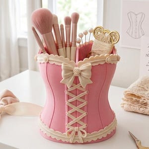 May include: A pink corset-shaped desk organiser featuring cream-coloured lace trim and a central bow detail. The organiser holds several makeup brushes and a spool of thread. It is accompanied by a matching pink scrunchie and gold-toned scissors.