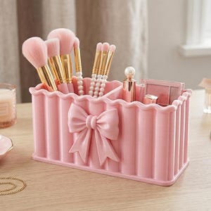 May include: A rectangular light pink makeup organiser featuring a scalloped edge and a prominent bow detail on the front. The organiser contains several makeup brushes with pink bristles and gold handles, a lipstick, and a small compact palette.