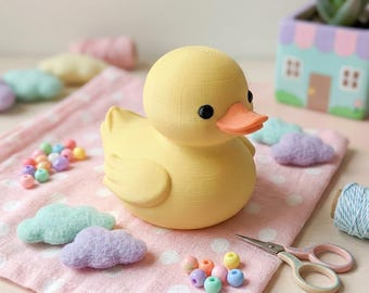 Puede incluir: Una figura de pato amarilla impresa en 3D con un cuerpo liso y redondeado, un pico naranja brillante y dos pequeños ojos negros. El pato está colocado sobre una tela rosa con lunares blancos, rodeado de pequeñas nubes de fieltro, cuentas de colores y tijeras.