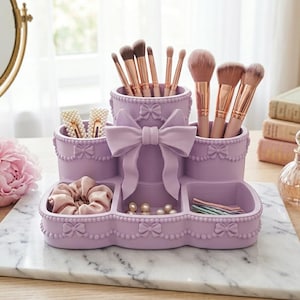 Op de afbeelding: Een lavendelkleurige bureau-organizer met een centraal verhoogd cilindrisch vak versierd met een grote strik, omringd door kleinere ronde en vierkante opbergvakken. De buitenkant is gedecoreerd met een kralenrand en kleine strikjes.