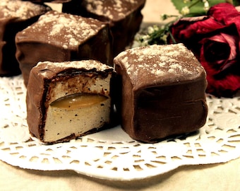 May include: A close-up of chocolate-covered marshmallow squares dusted with powdered sugar. The squares are arranged on a white doily. One square is cut open, revealing a caramel filling.