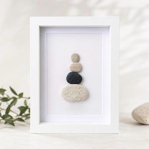 Pode incluir: Uma moldura retangular branca contendo uma pilha vertical de quatro pedras naturais lisas sobre um fundo branco liso. As pedras estão dispostas da menor para a maior, com três seixos bege claro e uma pedra cinza-escura na terceira posição.