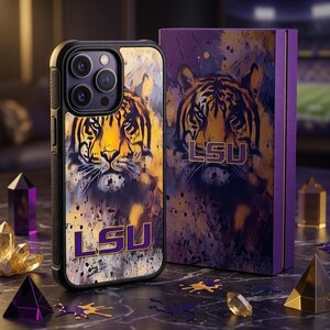 Puede incluir: Una funda de smartphone negra con un gráfico de cara de tigre con salpicaduras de pintura amarilla y morada y el texto LSU en morado. Al lado hay una caja rectangular morada a juego con el mismo gráfico de tigre y el texto LSU.