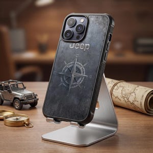 Könnte beinhalten: Eine dunkelgraue Smartphone-Hülle mit dem Jeep-Logo in silbernem Text oben und einer weißen Kompassrosengrafik in der Mitte. Die Hülle hat einen strukturierten schwarzen Rand und ist auf einem silbernen Metallständer montiert, mit robuster Optik.