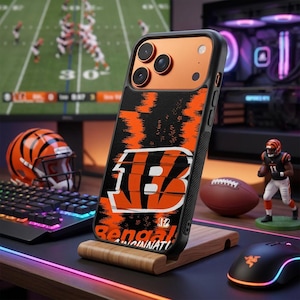 Puede incluir: Una funda para smartphone con un patrón de rayas de tigre en negro y naranja y un gran logotipo de la letra B en blanco y naranja. La funda incluye el texto Bengals Cincinnati en la parte inferior y tiene una zona de cámara naranja metálica.