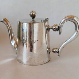 May include: A silver-toned metal teapot featuring a cylindrical body, a curved spout, and a C-shaped handle attached with spherical accents. The side of the teapot is engraved with the text "City Hotel" in a cursive script, and it includes a lid.
