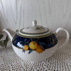 Può includere: Una teiera in ceramica bianca con una fascia blu scuro attorno al centro decorata con limoni gialli, foglie verdi e un bordo geometrico. La teiera include un coperchio abbinato con un piccolo pomello, un manico curvo e un beccuccio.