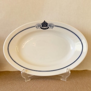 May include: An oval-shaped white ceramic platter featuring a dark blue double-line border around the rim. At the top center, there is a detailed dark blue crest or emblem with intricate scrollwork and a central shield design printed on the surface.