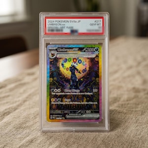Op de afbeelding: Een gegradeerde 2024 Pokemon SV8a JP Umbreon ex ruilkaart in een doorzichtige plastic houder. De kaart toont een donker Umbreon-personage omringd door kleurrijke zwevende edelstenen en een cirkelvormig patroon. Het label vermeldt 2024 POKEMON SV8a JP UMBREON ex #217 GEM MT.