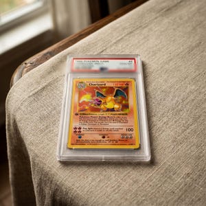 May include: A 1999 Pokemon Game Charizard trading card encased in a clear protective plastic slab. The card features a yellow border, a central illustration of the orange dragon-like creature, and red fire energy symbols with 120 HP text.