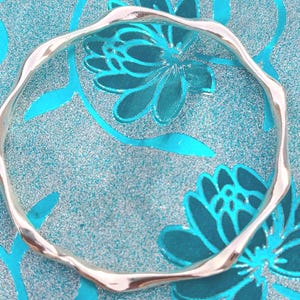 Può includere: Un bracciale rigido color argento con un design a bordo continuo, irregolare, ondulato o increspato. La superficie metallica è liscia e riflettente, formando una forma circolare che mantiene uno spessore costante lungo tutta la circonferenza.