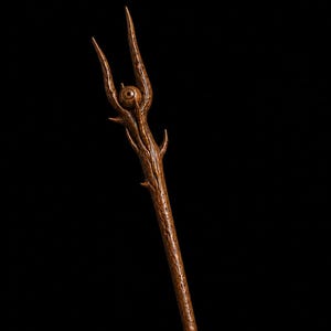 Op de afbeelding: Een bruine houten staf met een getextureerd, schorsachtig oppervlak en een drietandige top. Een realistisch, gedetailleerd oog is ingebed in het midden van de tanden, omringd door kleine, scherpe, doornachtige uitsteeksels langs de bovenkant.