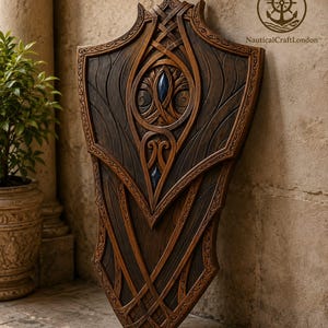 May include: A dark brown wooden shield featuring intricate carved patterns, layered sections, and two embedded dark blue gemstone accents. The shield displays the NauticalCraftLondon logo in the upper right corner, featuring a ship's wheel and anchor.