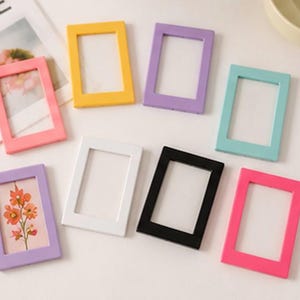 May include: A collection of eight rectangular picture frames in various solid colors including yellow, purple, light blue, pink, white, and black. One purple frame contains a small illustration of orange flowers on a light background.