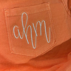 May include: A close-up of a bright orange pocket on a garment with the name "ahm" embroidered in white.