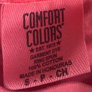 K&ouml;nnte beinhalten: Ein rosa Etikett mit dem Text "COMFORT COLORS" in Schwarz, gefolgt von "EST. 1975" mit einem Stern auf jeder Seite. Das Etikett enth&auml;lt auch den Text "GARMENT DYE", "RING SPUN", "100% COTTON", "MADE IN HONDURAS" und "S-P-CH" in Schwarz.