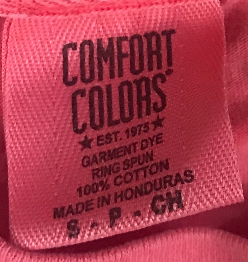 K&ouml;nnte beinhalten: Ein rosa Etikett mit dem Text "COMFORT COLORS" in Schwarz, gefolgt von "EST. 1975" mit einem Stern auf jeder Seite. Das Etikett enth&auml;lt auch den Text "GARMENT DYE", "RING SPUN", "100% COTTON", "MADE IN HONDURAS" und "S-P-CH" in Schwarz.