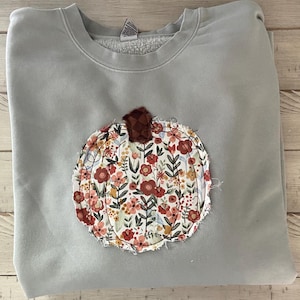 May include: A light gray sweatshirt with a floral pumpkin applique. The pumpkin is made of fabric with a pattern of red, pink, and yellow flowers and green leaves. The stem is a brown, textured fabric. The sweatshirt is folded on a light wood surface.
