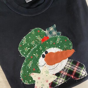 May include: Black sweatshirt with a snowman applique. The snowman has a green hat with a red berry, an orange carrot nose, and a plaid scarf. A white star with snowflakes is near the snowman's face.