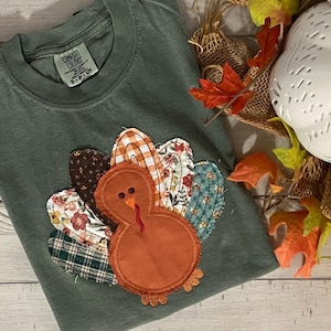 May include: A sage green Comfort Colors t-shirt featuring a felt turkey applique. The turkey has an orange body and a red wattle. The turkey's tail feathers are made of various patterned fabrics, including floral, plaid, and gingham.