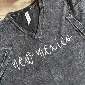 K&ouml;nnte beinhalten: Ein dunkelgraues V-Ausschnitt-T-Shirt mit dem wei&szlig;en, kursiven Text "new mexico". Das Shirt hat ein strukturiertes, ausgewaschenes Aussehen. Das Etikett lautet "Bella+Canvas".