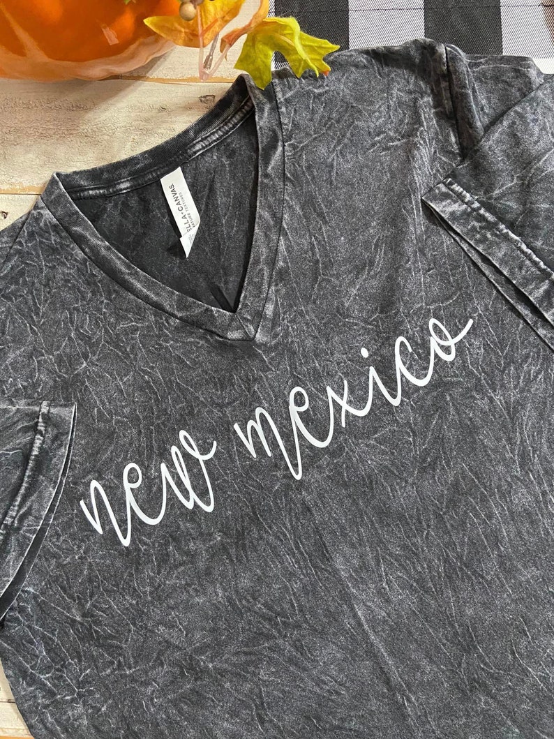 K&ouml;nnte beinhalten: Ein dunkelgraues V-Ausschnitt-T-Shirt mit dem wei&szlig;en, kursiven Text "new mexico". Das Shirt hat ein strukturiertes, ausgewaschenes Aussehen. Das Etikett lautet "Bella+Canvas".