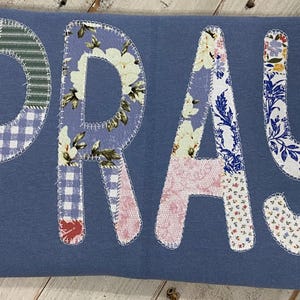 May include: A blue fabric item with the word "PRAY" appliquéd on it. The letters are made of various patterned fabrics, including floral, gingham, and striped designs. The letters are stitched onto the blue fabric.