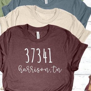 May include: A maroon short-sleeved t-shirt featuring the white text 37341 and harrison, tn in a handwritten font style. The shirt is layered with a beige t-shirt and a dark blue t-shirt, all featuring a crew neckline and a standard fit.