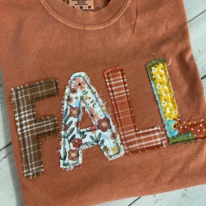 May include: A rust-colored t-shirt featuring the word FALL appliqued in fabric letters. The F is brown plaid, the A is a floral print, the L is orange plaid, and the second L is a yellow floral pattern with a dark red base. The letters have raw edges.